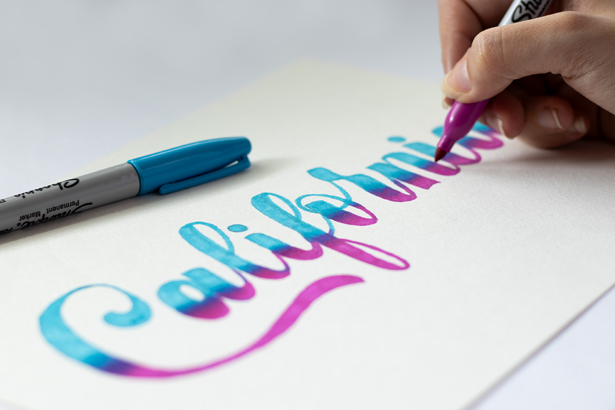 Person drawing script lettering that reads "California" in a blue and pink gradient.