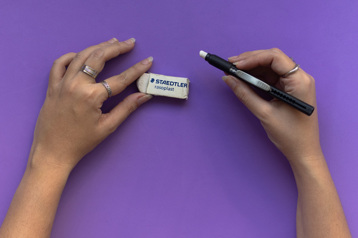 Two hands holding a Staedtler eraser (left) and a black retractable eraser (right) on a purple background.