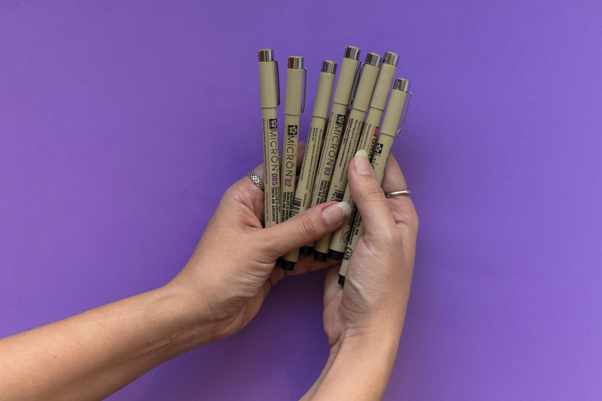 Hands holding seven Sakura Micron Pens of different pen tip sizes on a purple background.