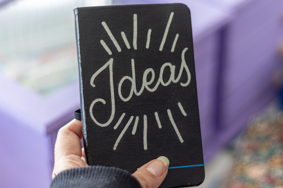 A hand holding a small black notebook. The notebook cover has a lettering in silver that reads "Ideas"