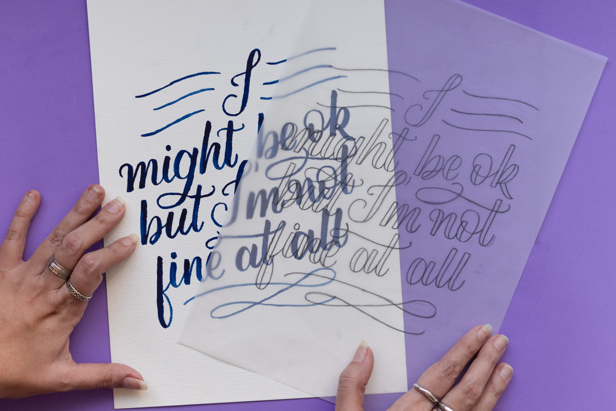 Hands holding two papers. On the left, a thicker paper with the words "I might be ok but I'm not fine at all" with a blue watercolor ink. On the left, the same words are written in pencil on a transparent tracing paper. The background is purple.