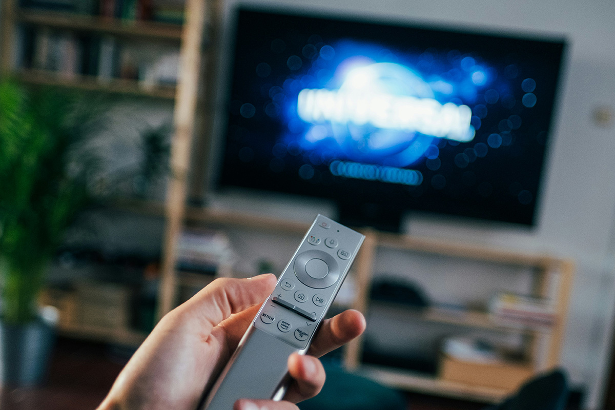 A hand with a silver remote control and a blurred TV in the background.