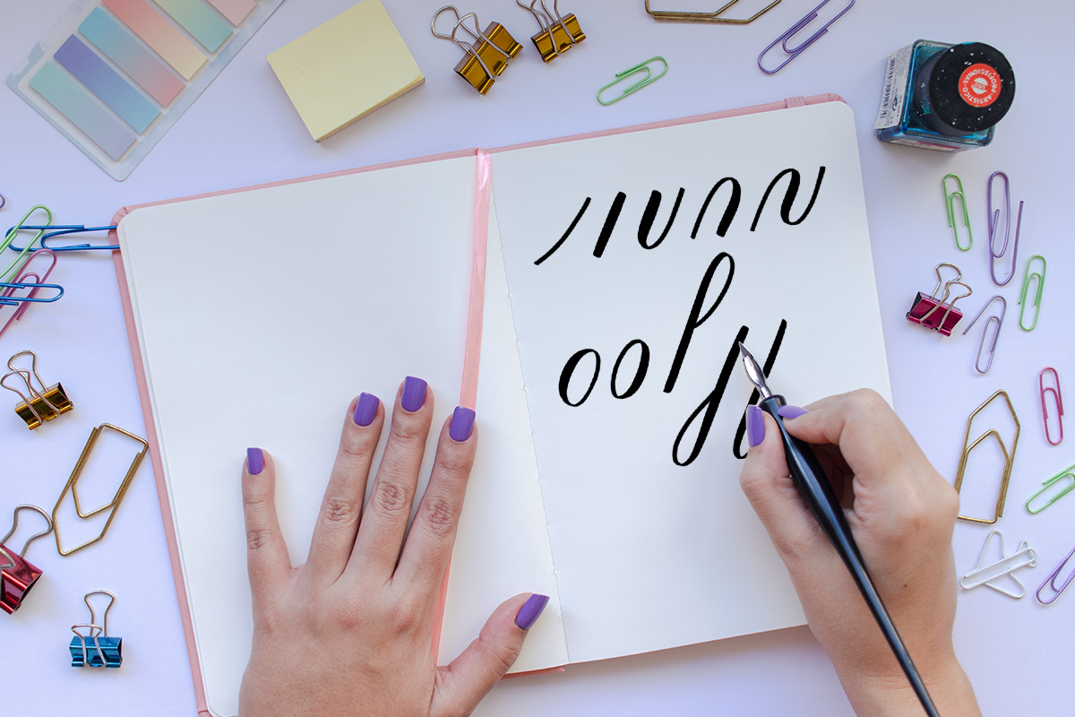 A notebook with a hand writing the calligraphy basic strokes with a dip pen, surrounded by clips, post-its, and ink.