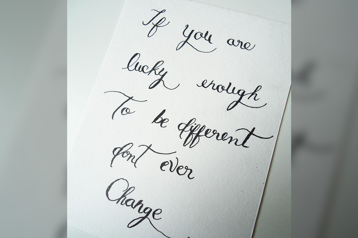 A paper with the words "If you are lucky enough to be different, don't ever change" in black script calligraphy.